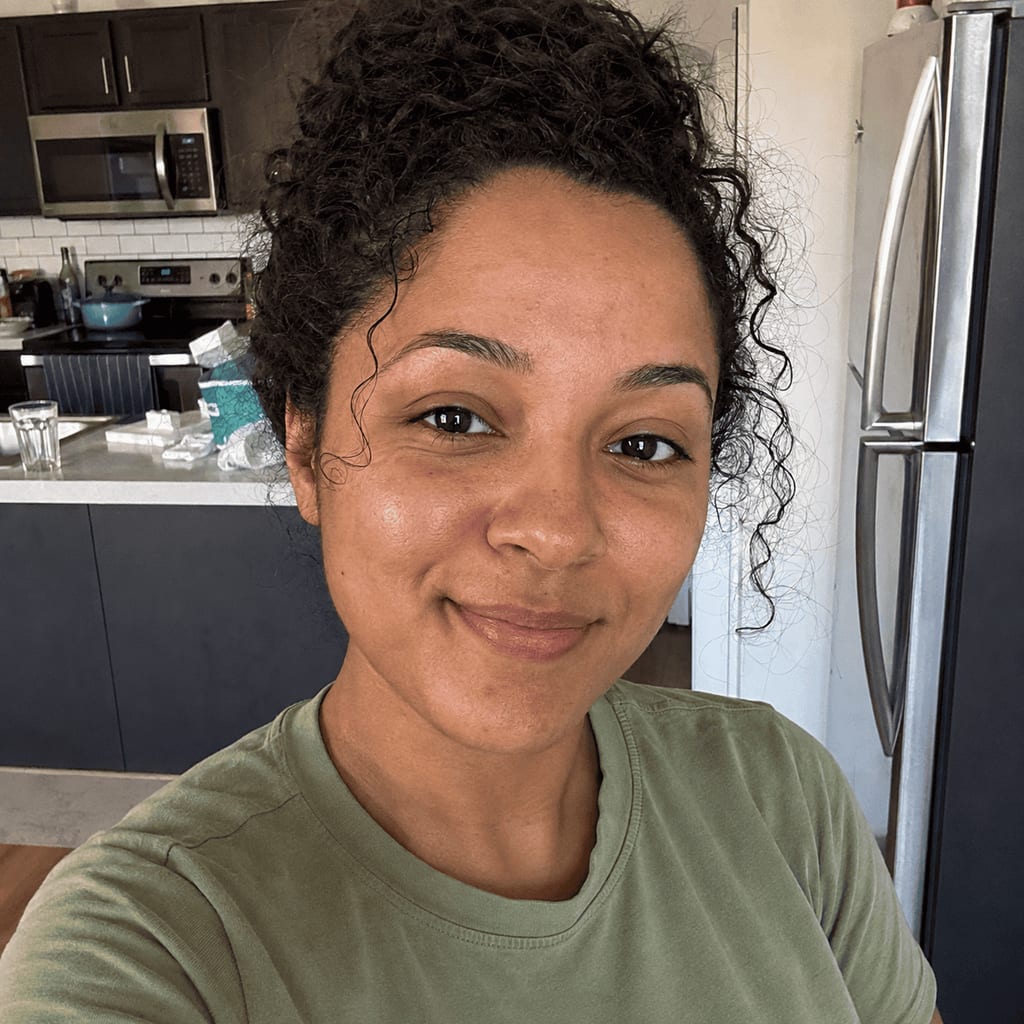 Woman with curly hair smiling in a kitchen, wearing a green t-shirt.