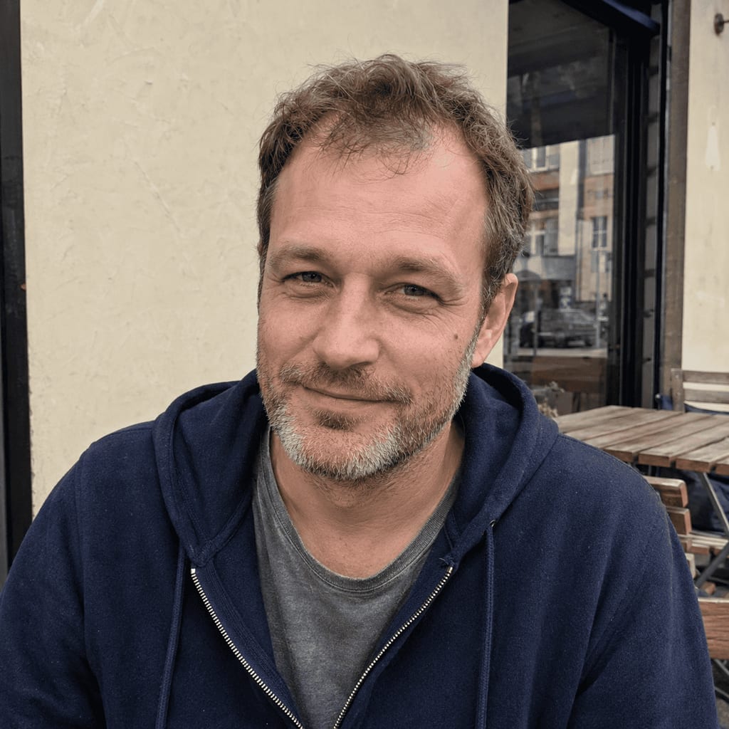Man with short greying hair and stubble, wearing a navy hoodie, smiling outside a cafe